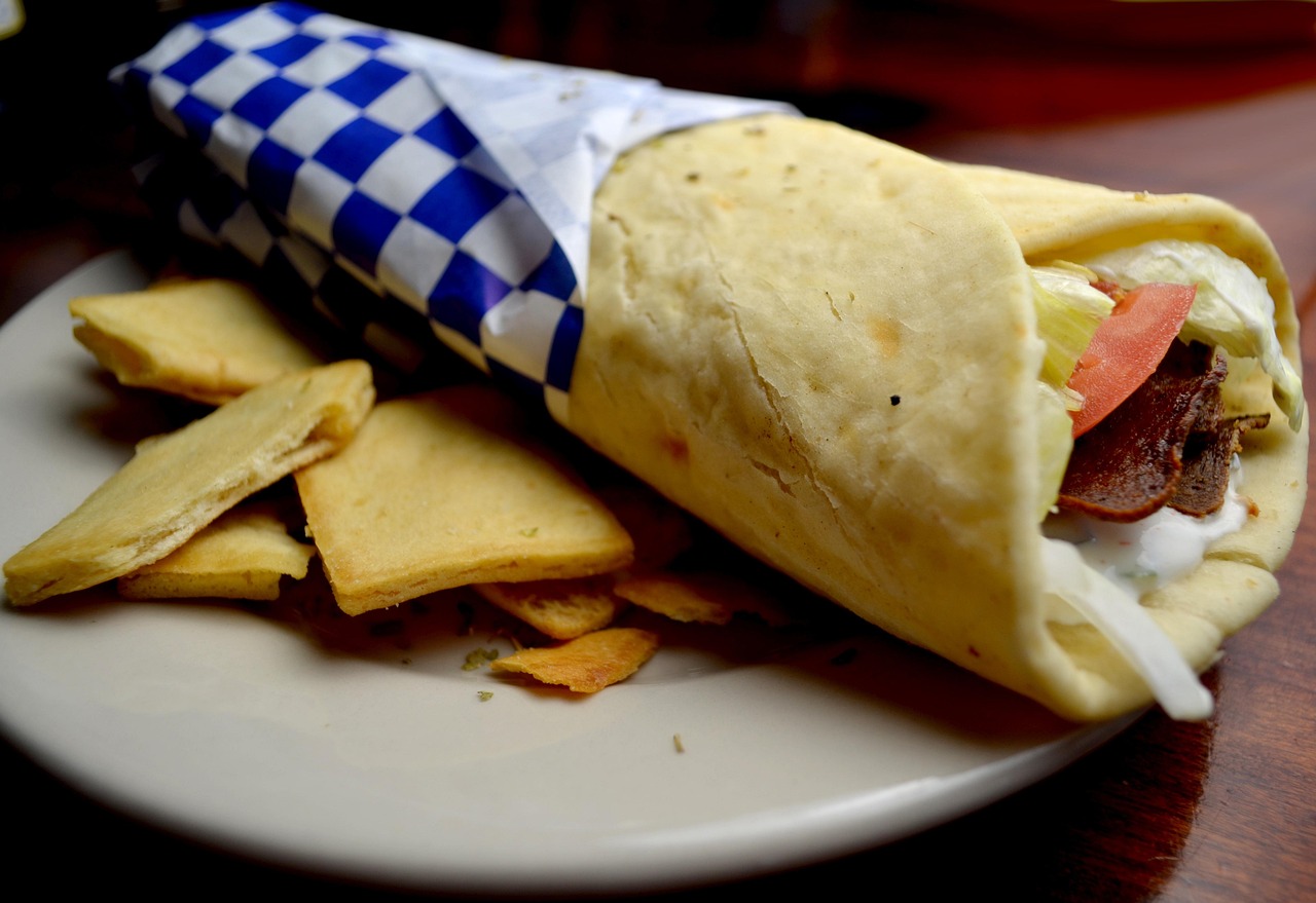 A gyro wrapped in blue checked paper, with lettuce, tomato, meat and white sauce, beside pita chips on a plate
